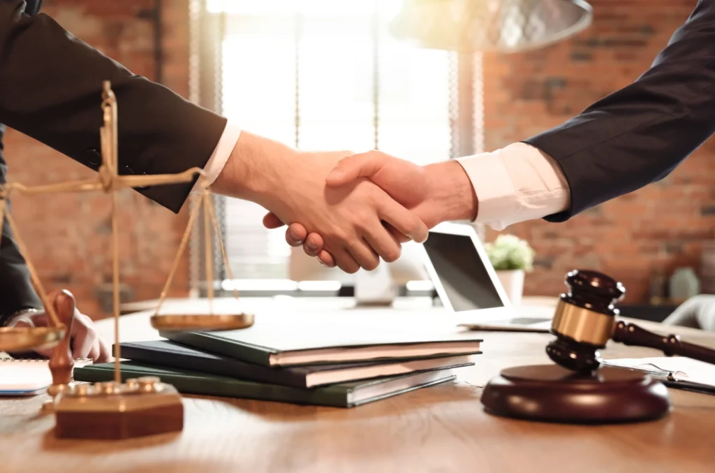 Two people in business suits shake hands across a desk in a law office, with a gavel, legal scales, and stacked documents visible in the foreground.